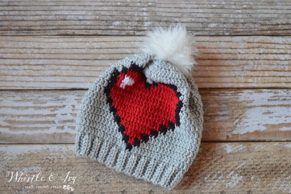 crochet hat with red pixelated heart graph on the front, with white fur pom-pom