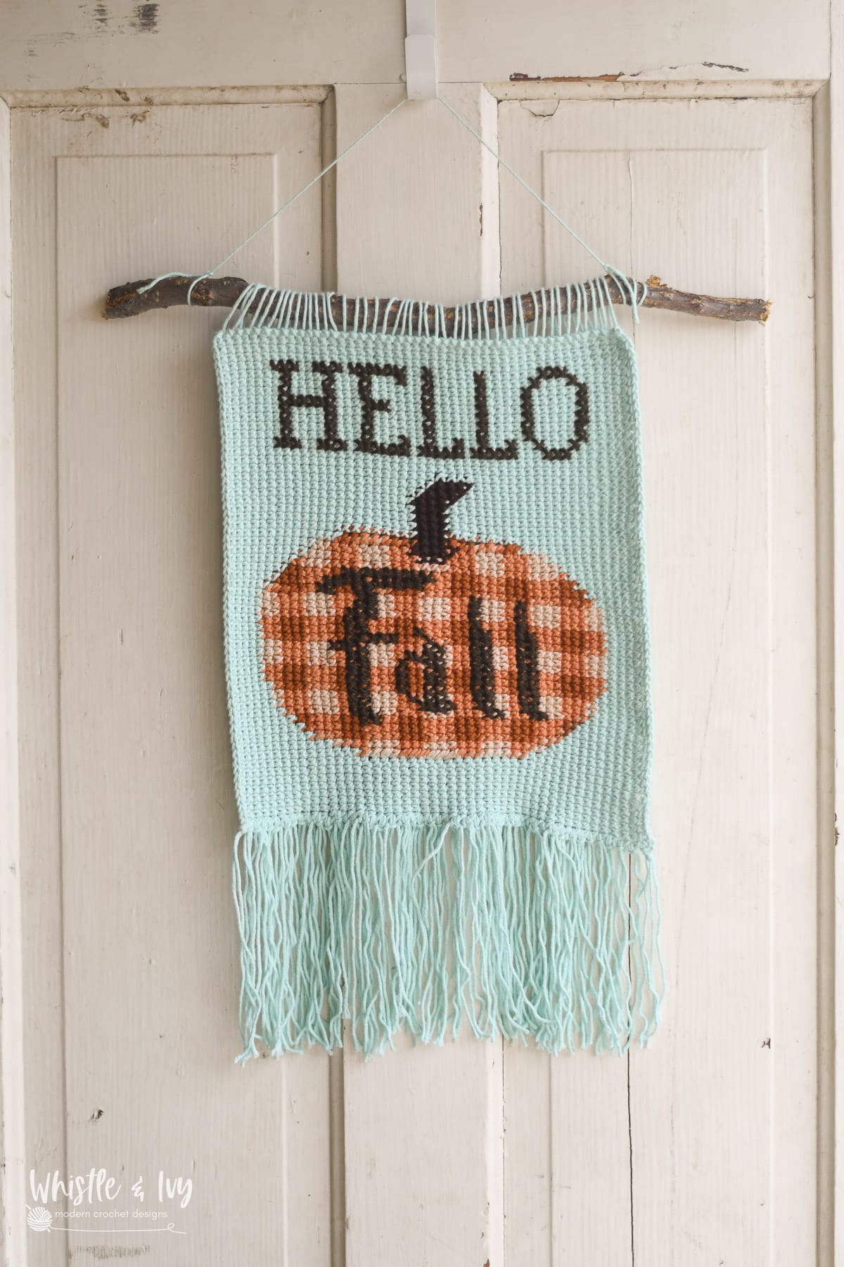 Crochet pumpkin wall hanging with orange gingham color work with Hello Fall stitched in cross stitch on the front with fringe across the bottom